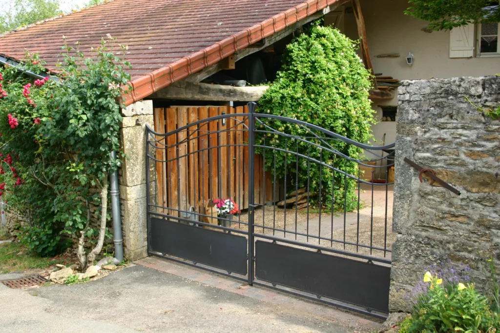 Vue du portail à barreaux arrondi intégré dans son environnement d'entrée de propriété. La cohérence entre le portail, la clôture et le pilier de maçonnerie existant est étudiée dès la conception pour un résultat harmonieux et valorisant. Chaque projet est précédé d'un relevé sur site et d'un dessin soumis au client pour validation avant fabrication.