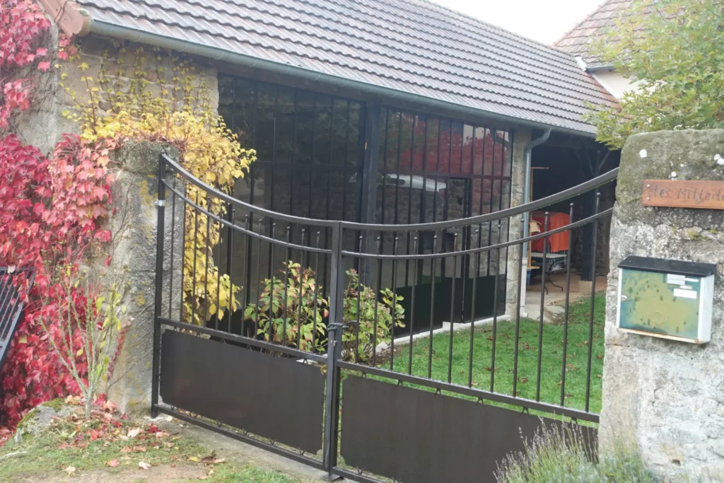 Un portail deux battants en fer forgé noir avec : Barreaux verticaux droits sur toute la hauteur Courbe concave en partie haute — le dessus s'incurve vers le bas en arc rentrant Soubassement plein sur les deux vantaux en partie basse Petits embouts décoratifs en tête de barreaux Piliers en pierre de taille anciens encadrant le portail En arrière-plan : une verrière noire type atelier couvrant une galerie