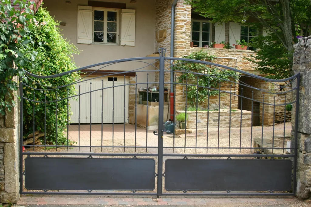 Vue du portail à barreaux arrondi intégré dans son environnement d'entrée de propriété. La cohérence entre le portail, la clôture et le pilier de maçonnerie existant est étudiée dès la conception pour un résultat harmonieux et valorisant. Chaque projet est précédé d'un relevé sur site et d'un dessin soumis au client pour validation avant fabrication.